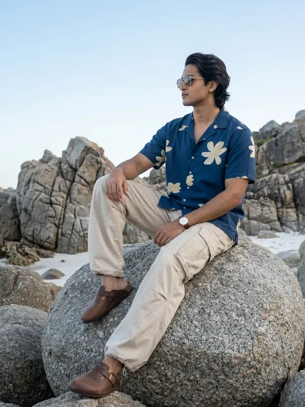 AI-generated menswear image of a man in a navy floral shirt and cargo trousers seated on a beach boulder